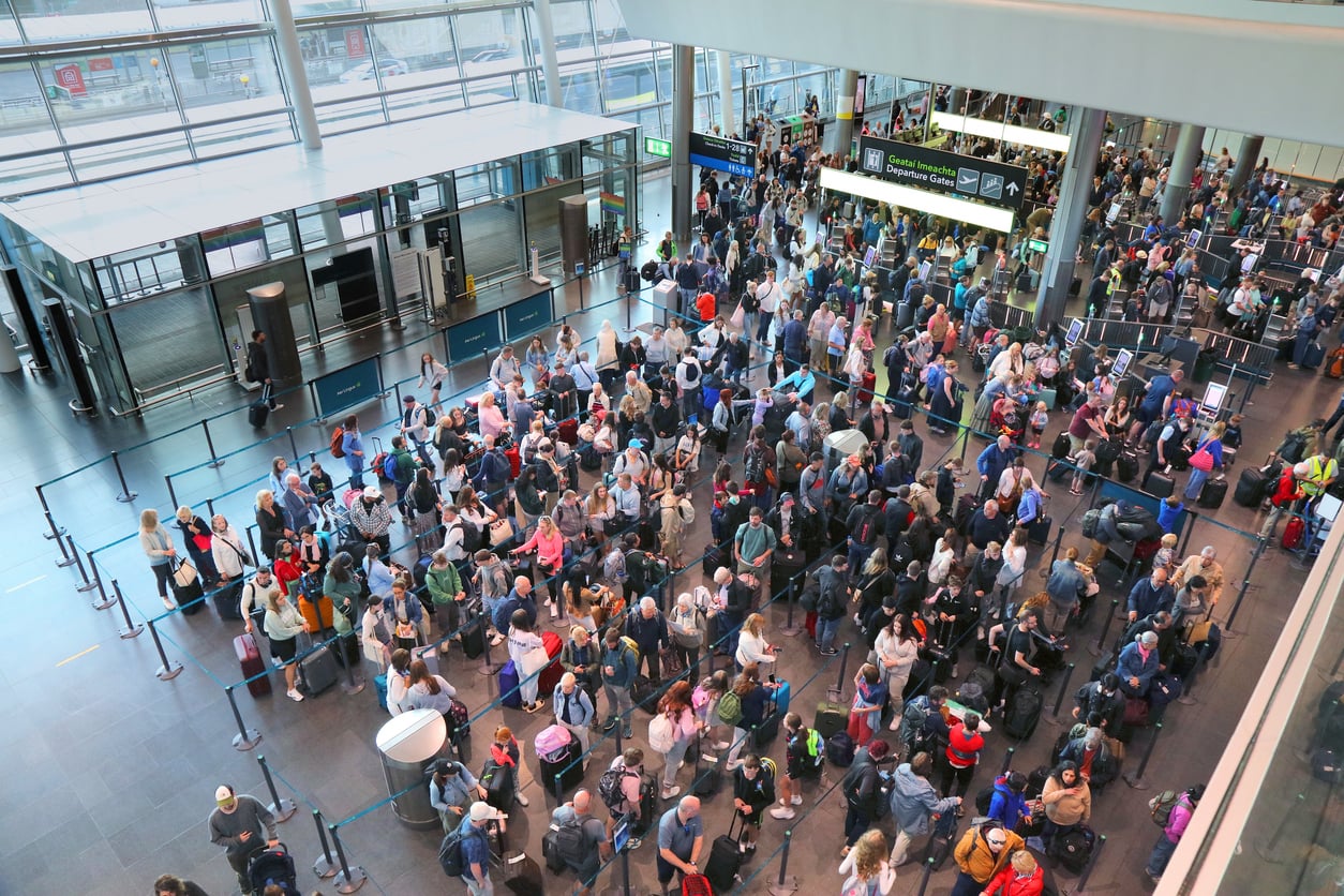 A large crowd of travelers with luggage waits in long lines at an airport check-in area, under a high glass ceiling.