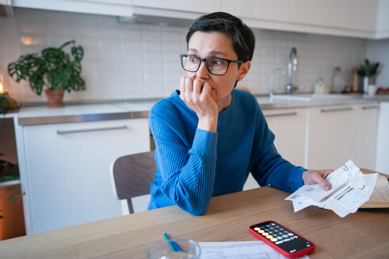 A person wearing glasses sits at a kitchen table, looking worried while holding bills. A smartphone with a calculator app and papers are on the table in front of them.