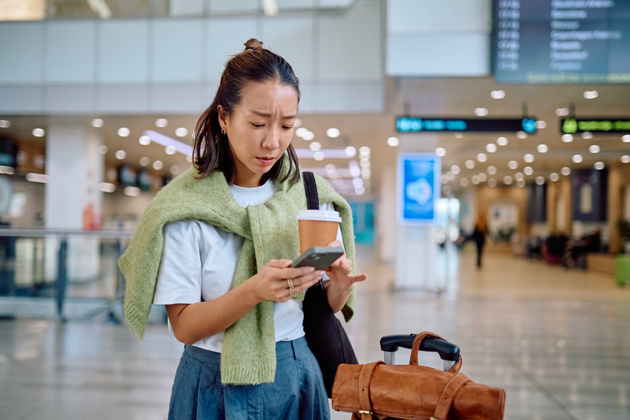 A woman stands in an airport holding a coffee cup and looking at her phone with a concerned expression. She has a sweater draped over her shoulders and her luggage beside her. Flight information screens are visible in the background.