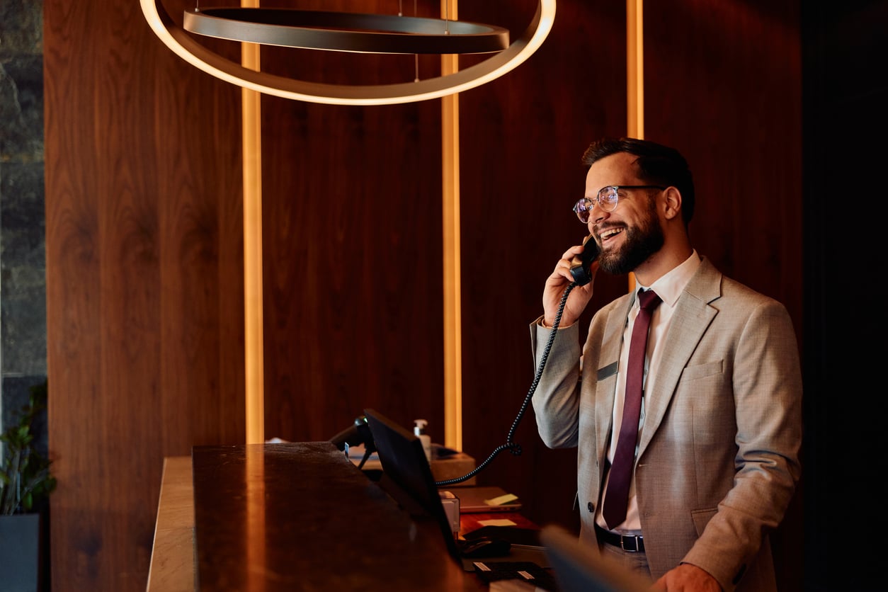 A man in a suit stands at a reception desk, smiling while talking on the phone. Warm lighting and wood paneling create an inviting, modern atmosphere that subtly recalls the charm of things hotels used to offer guests.