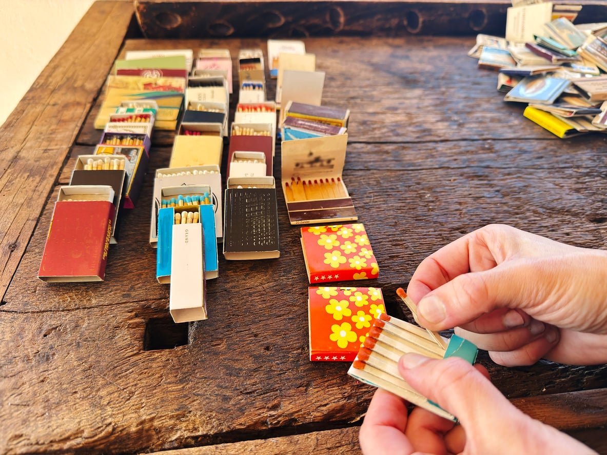 A person holds an open matchbox with matches, while more colorful matchboxes—reminiscent of the things hotels used to offer guests—are neatly arranged on a rustic wooden table in the background.