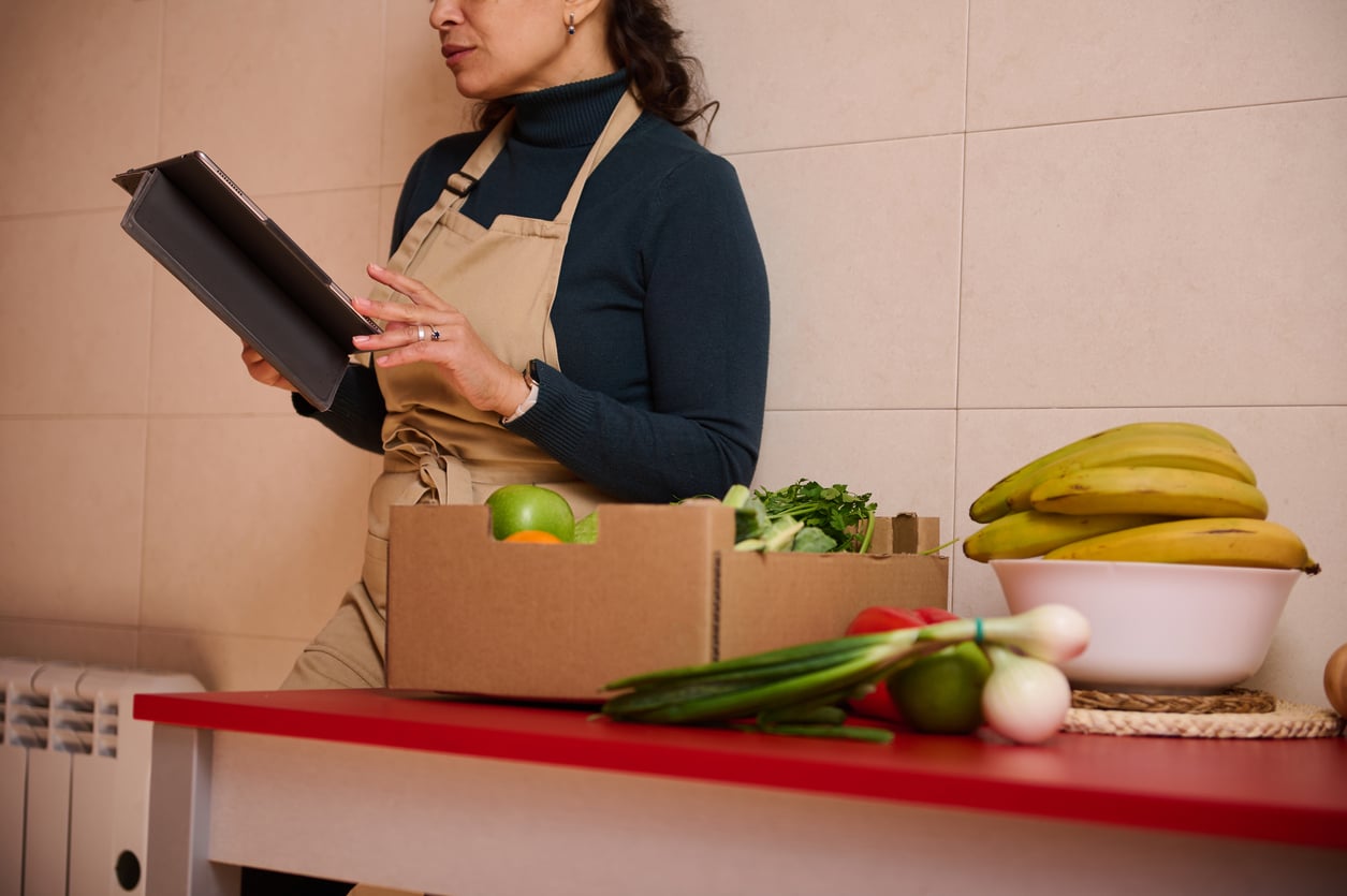 A woman in an apron holds a tablet and stands by a table with a box of fresh produce and a bowl of bananas, preparing to cook.