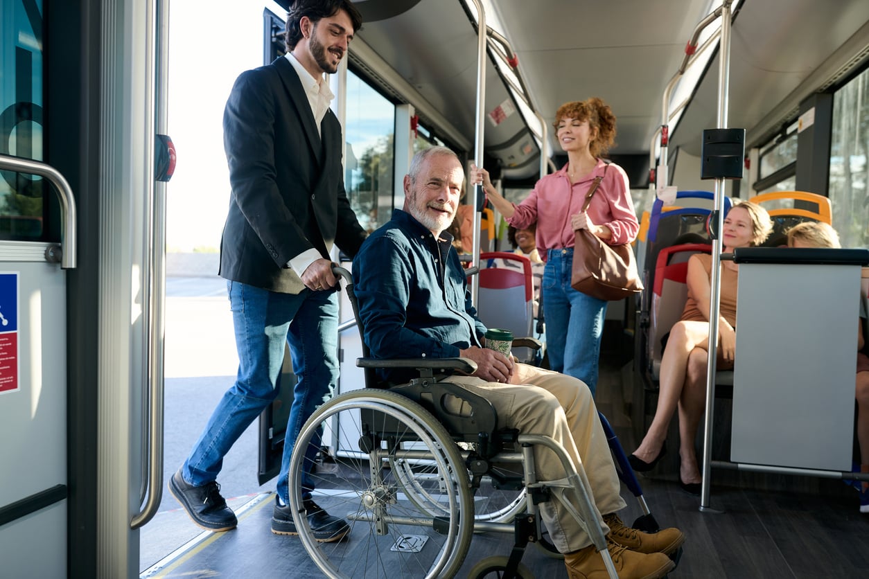 A man in a wheelchair is being assisted onto a bus by another man, while two women inside the bus smile and watch, reflecting inclusive travel behaviors that make public transportation accessible to everyone.