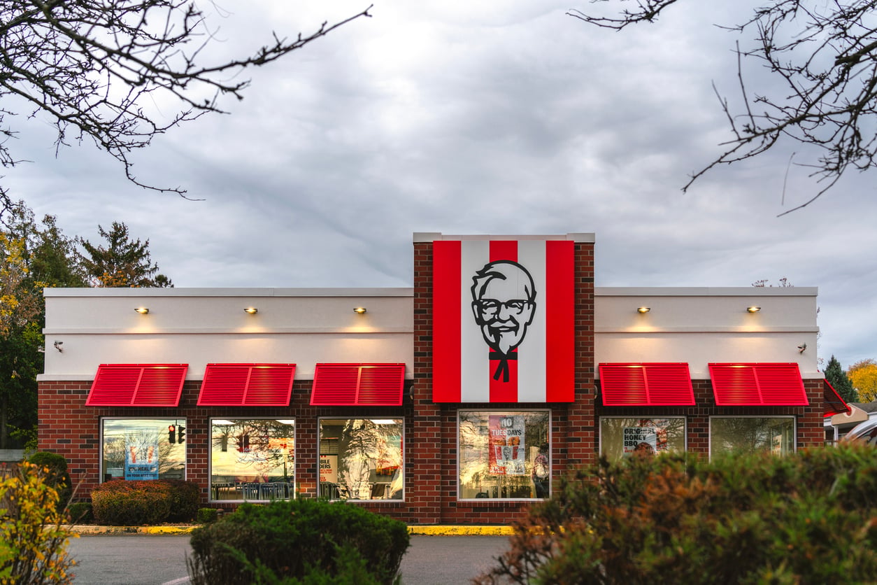 A KFC restaurant with red and white decor, large Colonel Sanders logo on the building, red awnings over the windows, and a cloudy sky in the background. Shrubs frame the foreground—setting the scene for where countless Wild Fast-Food Facts come to life.
