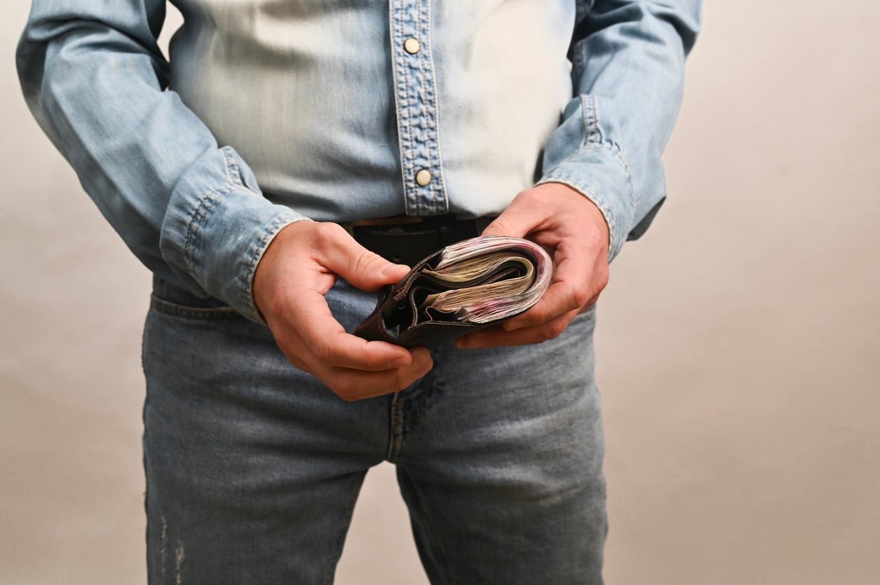 A person wearing a denim shirt and jeans holds an open wallet filled with cash, showing several banknotes inside. The individual’s face is not visible in the image.