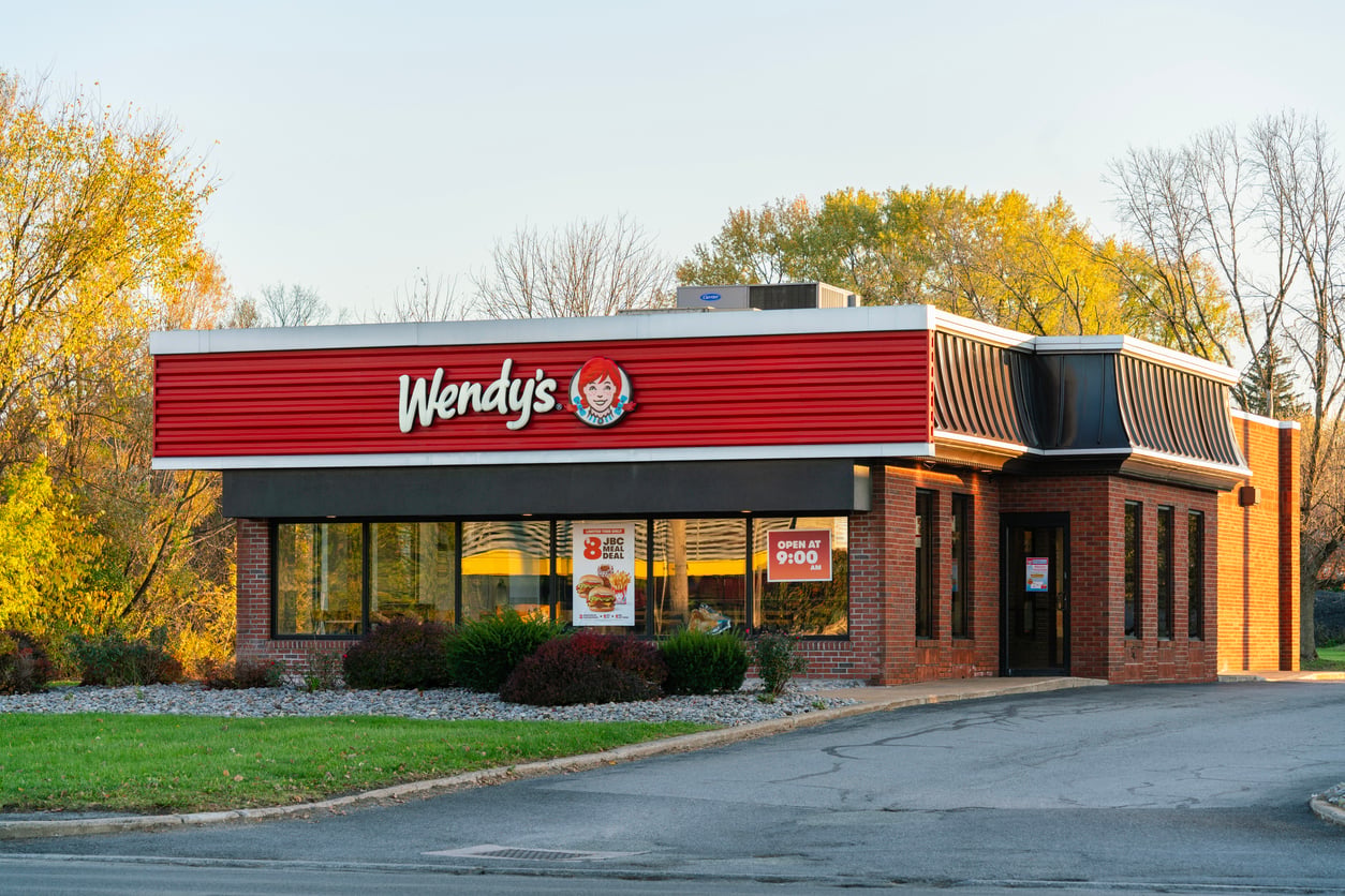 A Wendy’s fast-food restaurant with a red and white sign, surrounded by trees and bushes, under a clear blue sky. The building is brick with large windows and a drive-thru lane.