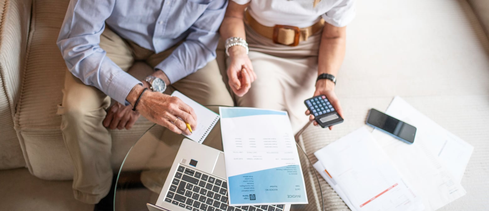 Two people sit on a couch working on finances with a laptop, paperwork, a calculator, and a notebook on the table in front of them. As one points at the notebook—possibly noting their travel habits—the other holds a calculator, helping organize expenses and plan ahead.