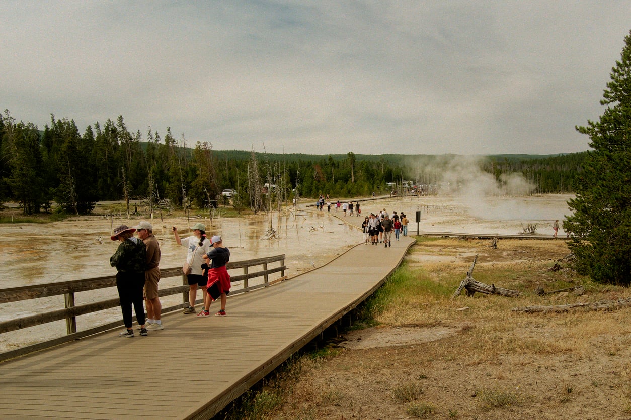 People walk along a wooden boardwalk through a geothermal area with steam rising, surrounded by trees and grassy terrain under a cloudy sky.