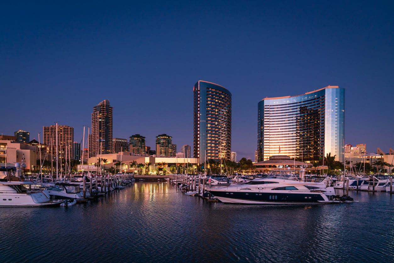Marina with docked boats in the foreground, modern high-rise buildings and city skyline illuminated at dusk in the background, calm water reflecting lights—a serene setting perfect for relaxation and reflection after age 50.