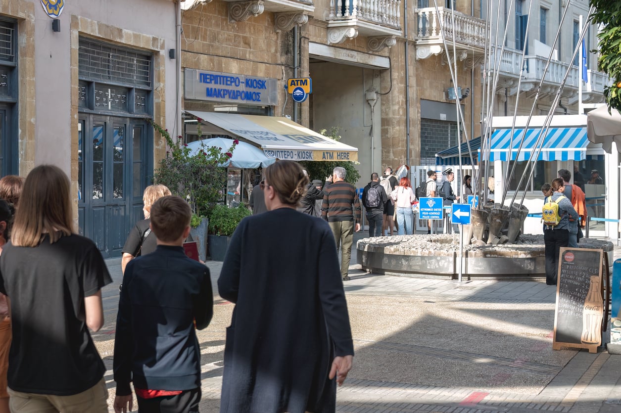 People walk near a fountain and kiosk on a sunny street, with a blue ATM sign and various shops in the background. Outdoor seating and a chalkboard menu are visible, serving as a vibrant meeting spot for locals and visitors alike—though some may notice signs reminding travelers of current travel restrictions and advisories in the area.