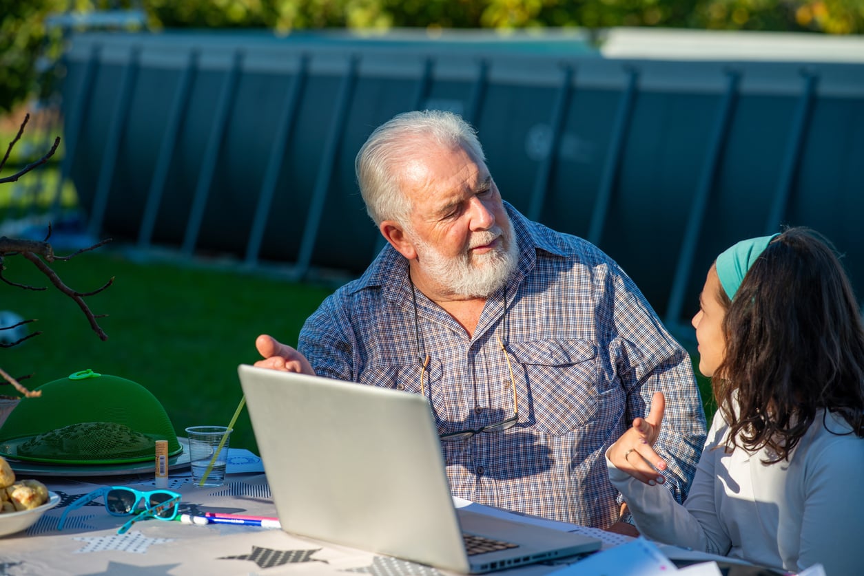 An older man and a young girl sit at an outdoor table with a laptop, talking and gesturing to each other on a sunny day. A swimming pool and green lawn are in the background.