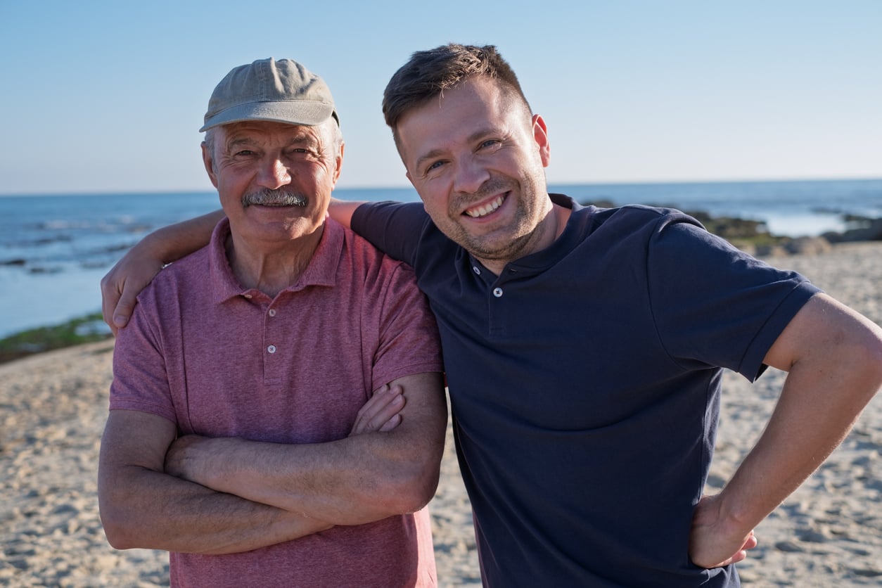 Two men stand on a sandy beach with the ocean behind them. The older man wears a cap and a pink shirt, while the younger man in a navy shirt has his arm around the older man’s shoulder. Both are smiling at the camera.