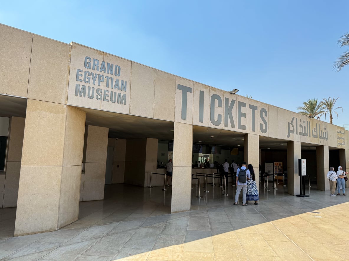 People gather at the ticket counter outside the Grand Egyptian Museum. The building has large signs in English and Arabic and is under a clear blue sky.