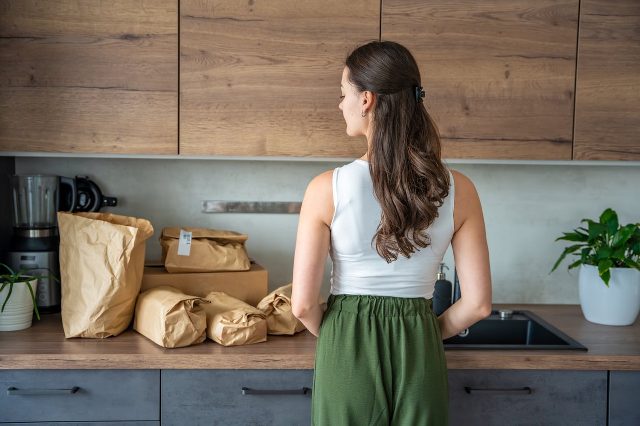 A woman with long brown hair, wearing a white top and green pants, stands in a kitchen and looks at several grocery bags placed on the counter.