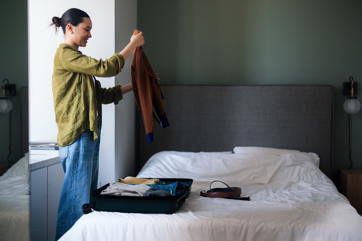 A woman stands by a bed, smiling as she packs clothes into an open suitcase. The room has a neatly made bed, soft lighting, and a calm atmosphere.