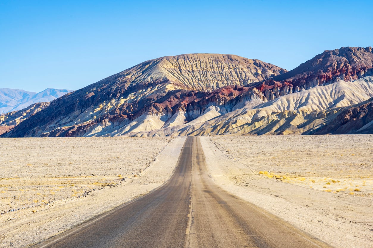 A straight road leads through a dry desert landscape toward colorful, rocky hills under a clear blue sky—an iconic scene that perfectly captures some classic Travel Facts About The United States.