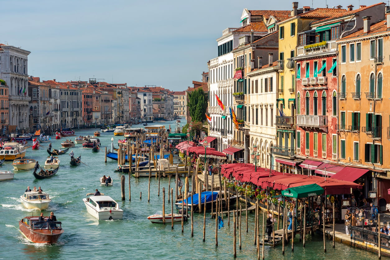 Colorful buildings line the Grand Canal in Venice, Italy. Boats and gondolas travel on the water, while people sit under red umbrellas at a waterside café, perhaps debating whether to risk the notoriously bad food at some tourist spots. Wooden posts stick out of the canal near the busy promenade.