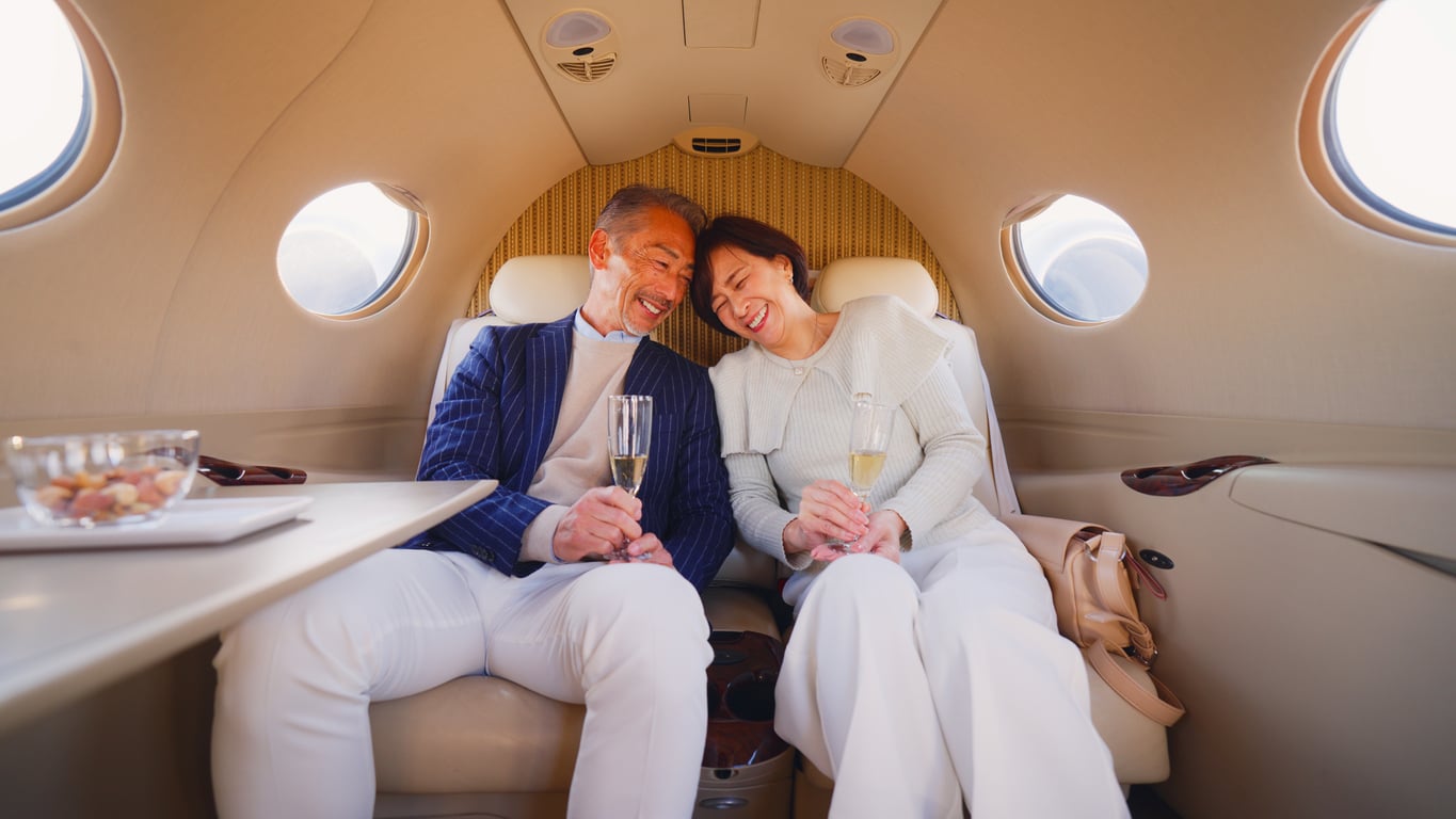 An older couple, dressed elegantly in white and sitting close together, smile and clink glasses of champagne while relaxing in the luxurious cabin of a private jet, showcasing their refined travel habits.