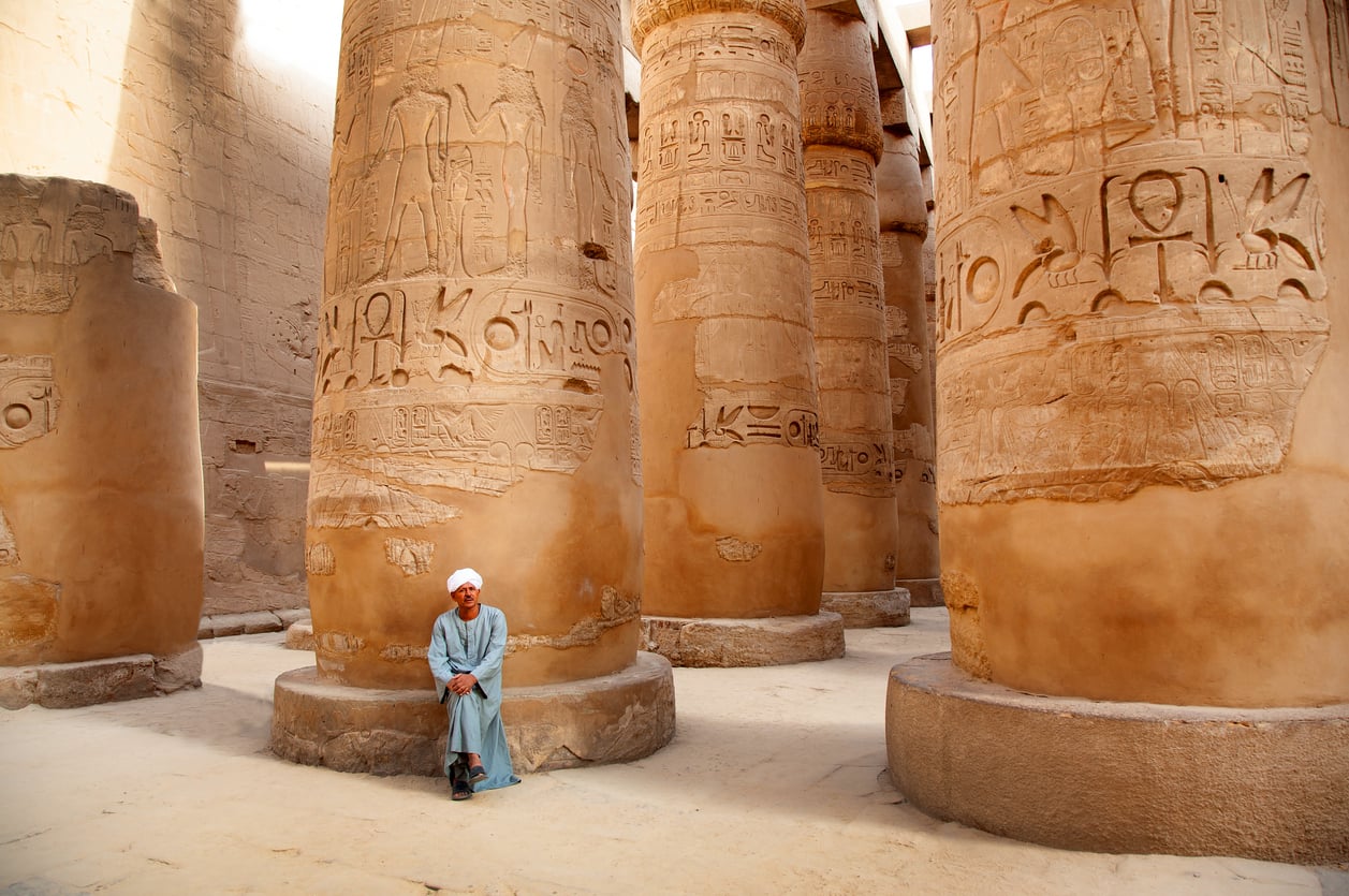 An elderly person in blue traditional clothing sits on the base of a large ancient Egyptian column with hieroglyphics, surrounded by several similar columns in sunlight.