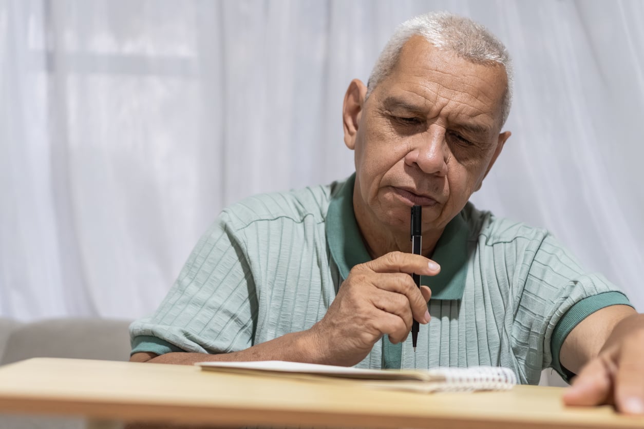 Older man with short gray hair, wearing a light green polo shirt, sits at a table thoughtfully holding a pen to his chin and looking at an open notebook in front of him.