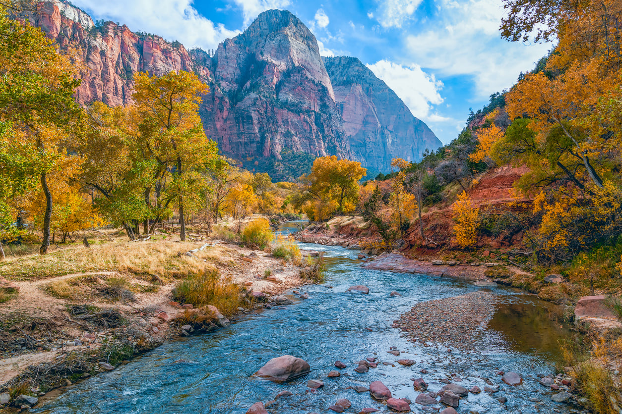 A river flows through a scenic valley with autumn trees, red rock cliffs, and mountains under a blue sky with scattered clouds, making it one of the most picturesque vacation spots for nature lovers.