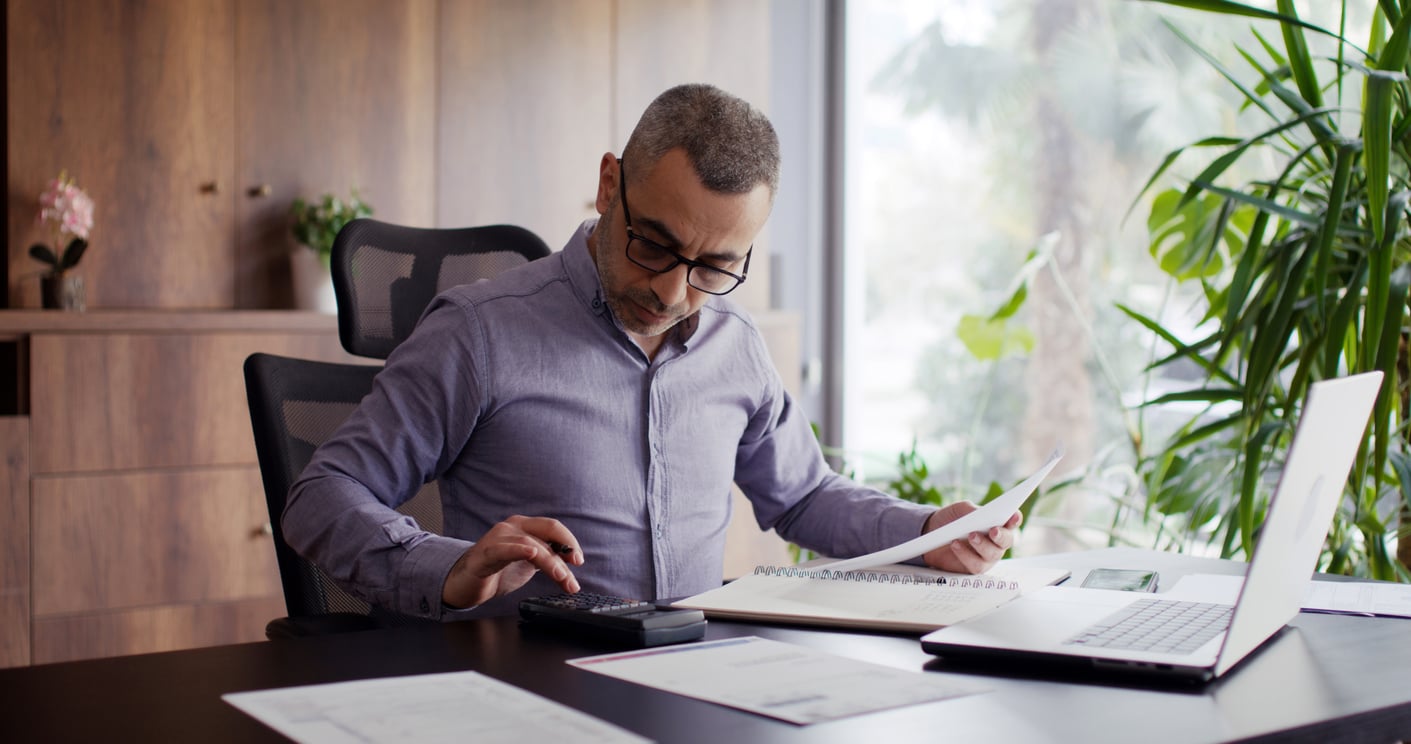 Man with glasses sits at a desk, using a calculator and holding papers. A laptop is open in front of him, and the room has plants and wooden shelves.