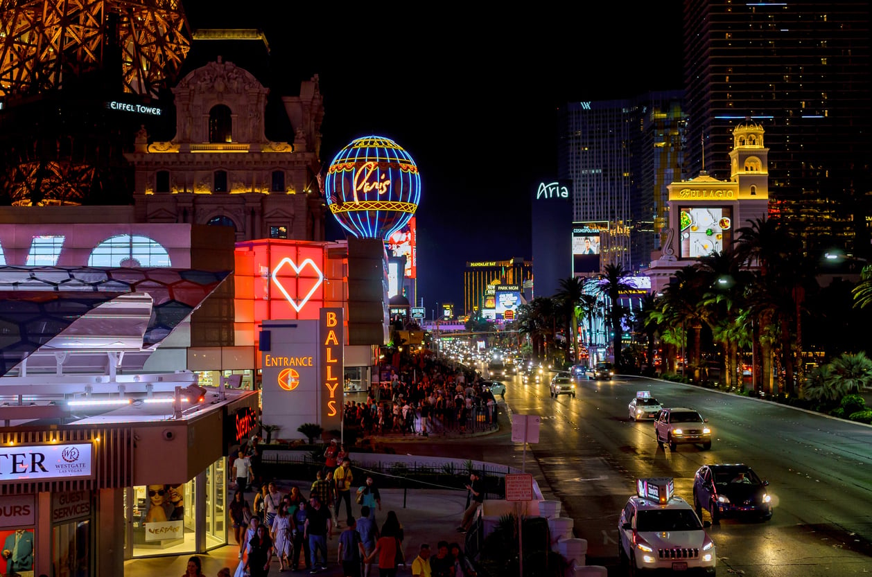 Las Vegas Strip at night with bright neon lights, crowds walking on sidewalks, and cars driving on the busy street, featuring Bally’s and Paris Las Vegas signs.