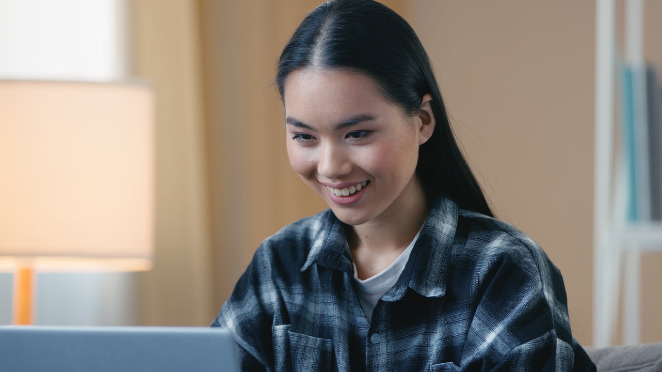A woman with long dark hair and a plaid shirt smiles while looking at a laptop screen in a cozy, softly lit room.