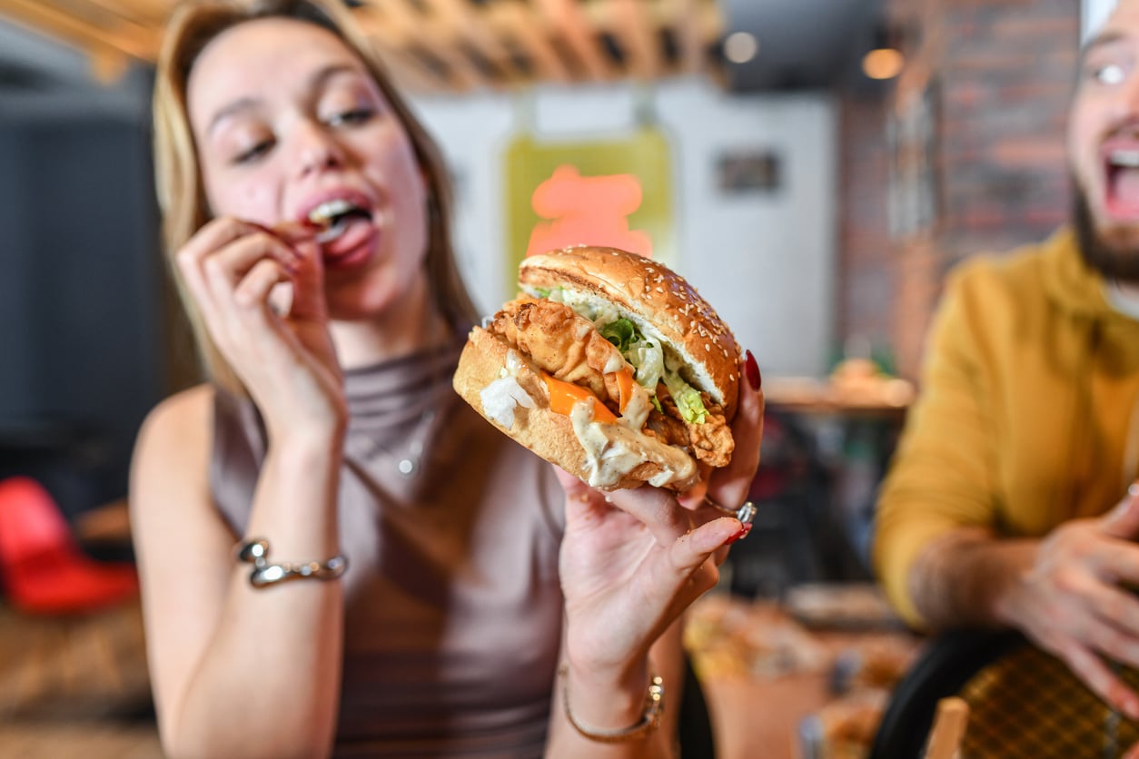 A woman holds a large fried chicken sandwich with lettuce and tomato, smiling and about to eat, while a man in the background laughs.
