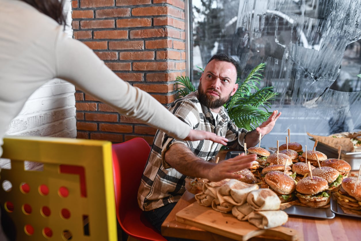 A man sitting at a table filled with burgers and wraps looks surprised and raises his hands defensively as a person reaches toward him.