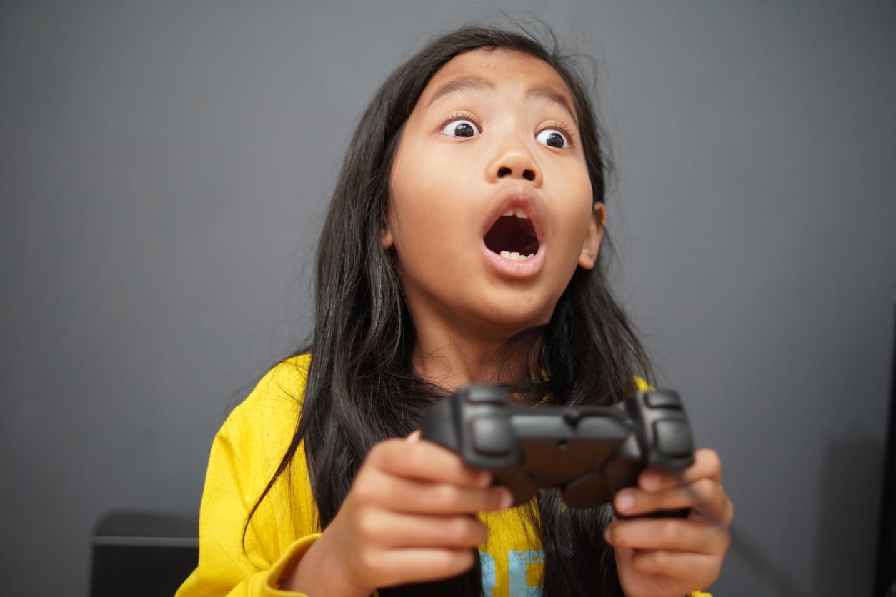 A young person with long dark hair, wearing a yellow shirt, holds a game controller and looks up with a surprised, wide-eyed, and open-mouthed expression against a plain gray background.