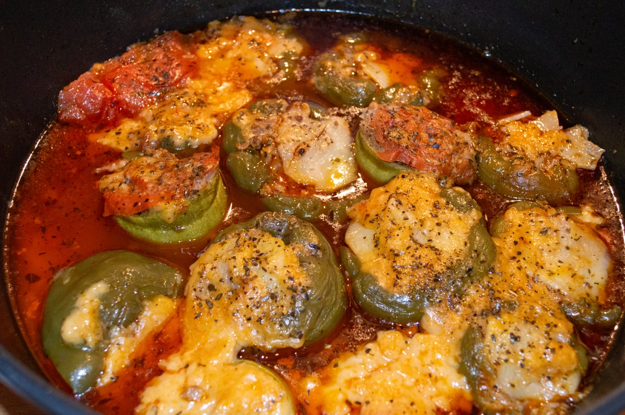 Stuffed green bell peppers topped with melted cheese and pepper, cooked in a tomato-based sauce inside a black pot.