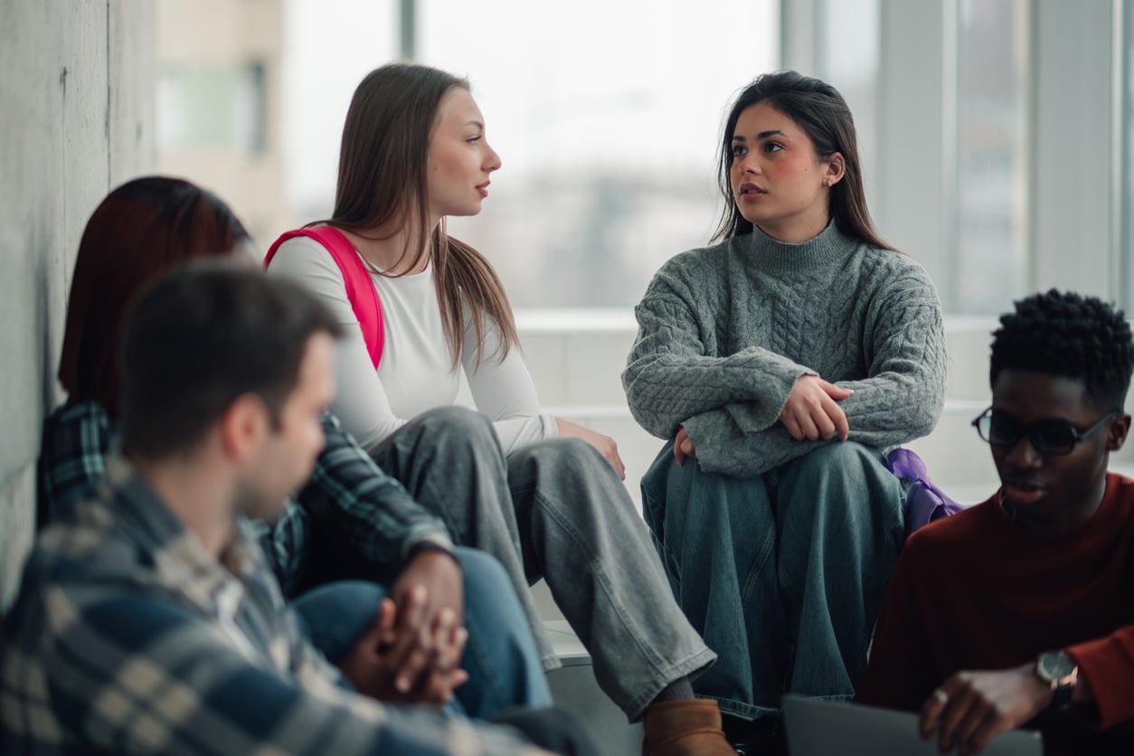 A group of young adults sit together indoors, talking and listening. Two women in the center appear engaged in conversation, while others sit nearby. Natural light comes in through large windows behind them.