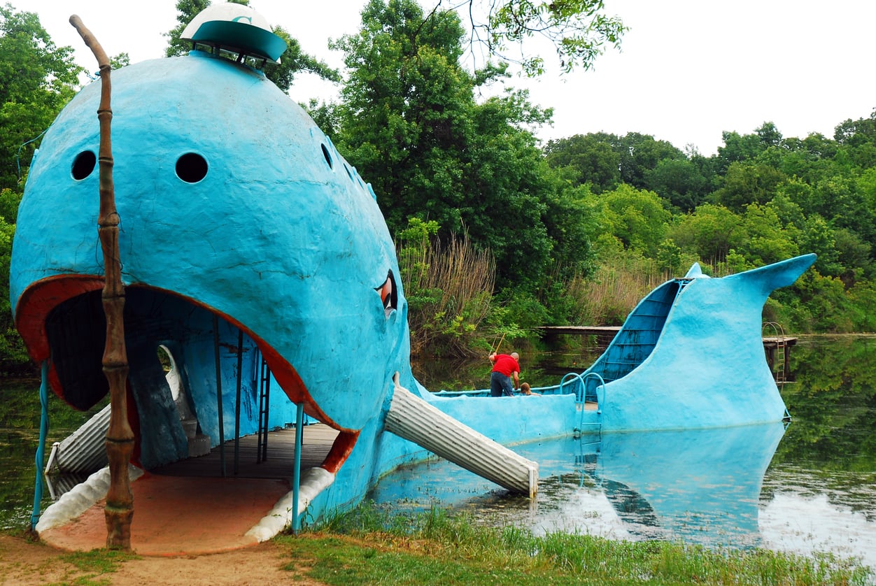 A large blue whale-shaped structure sits partly in a pond, surrounded by trees. The whale has an open mouth, a slide, and a dock, with a person in red nearby.