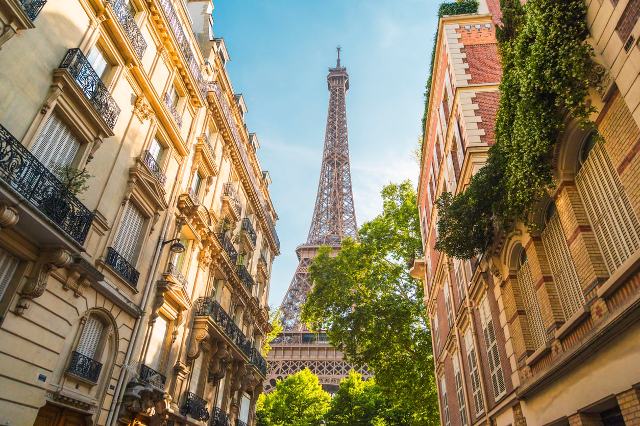 The Eiffel Tower rises between two classic Parisian apartment buildings on a sunny day, with green trees and a clear blue sky in the background—a picturesque scene that offers little hint of the bad food experiences some travelers report in nearby tourist cafés.