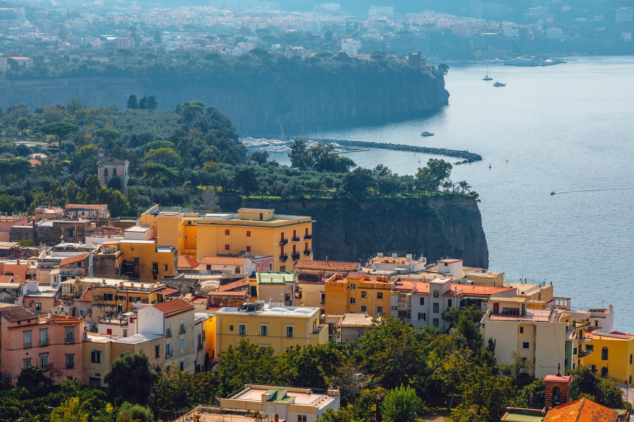 Colorful buildings with orange rooftops overlook a cliffside by the sea, with green trees and a distant coastline visible under a hazy sky. Sailboats are anchored near the shore.