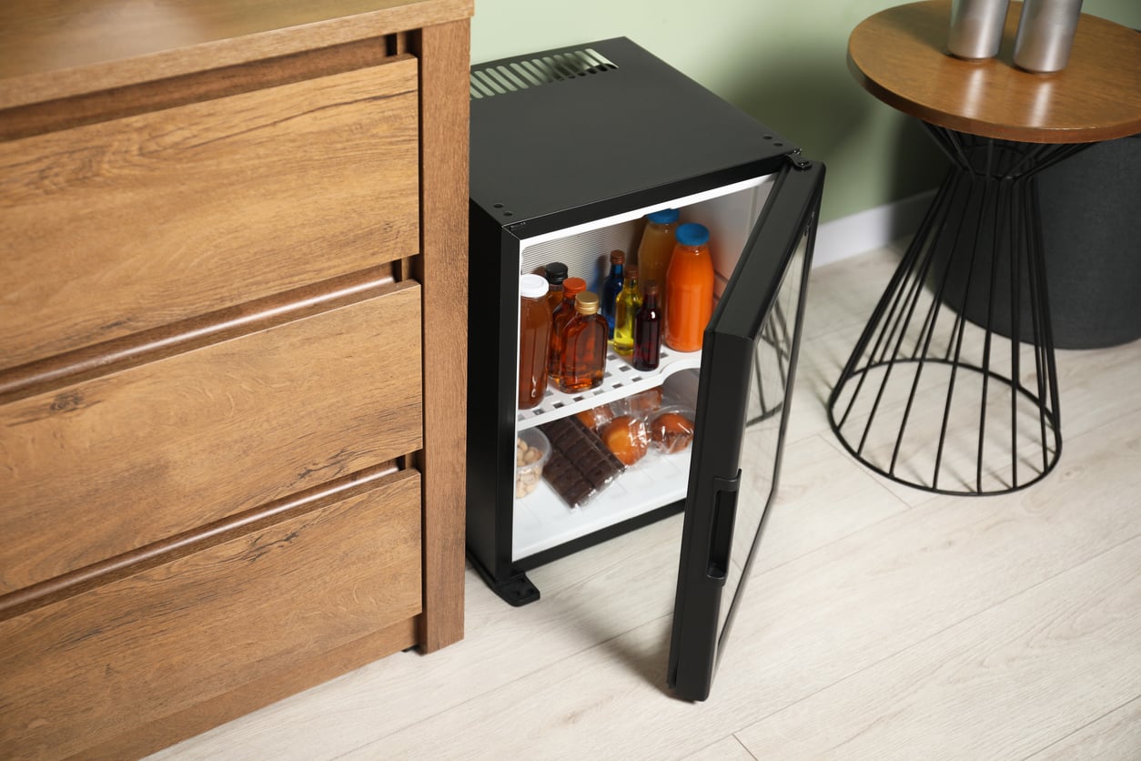 A small black mini fridge with an open door sits on the floor next to a wooden dresser and round table, reminiscent of the things hotels used to offer guests, showing shelves with bottles and drinks inside.