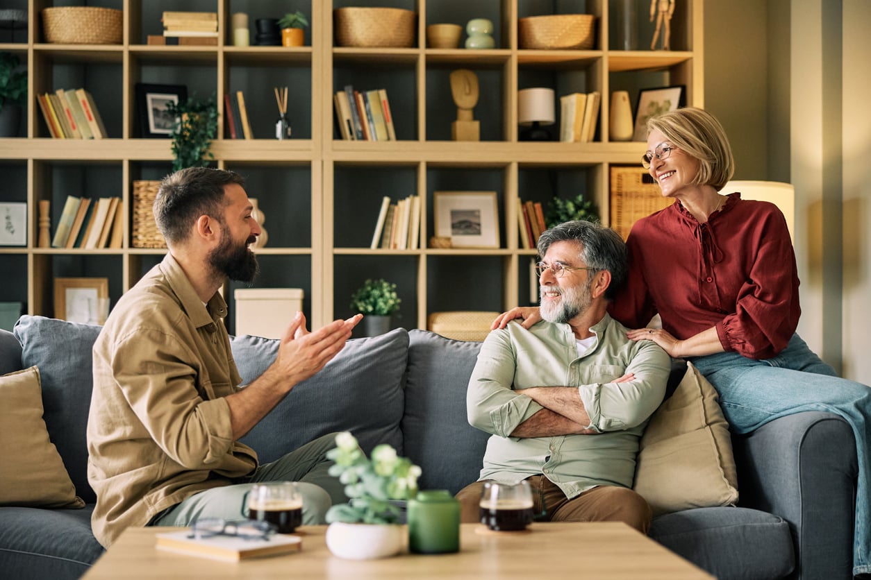 Three adults—a younger man and an older couple—are smiling and talking together in a cozy living room with a large bookshelf in the background, reflecting on Baby Boomer changes that have shaped their lives and relationships.
