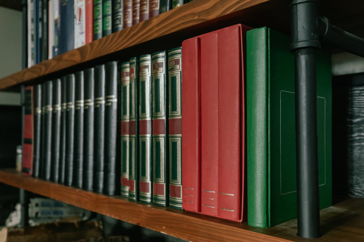 A close-up of a wooden bookshelf filled with green, red, and black hardcover books and binders, next to a black metal pipe.