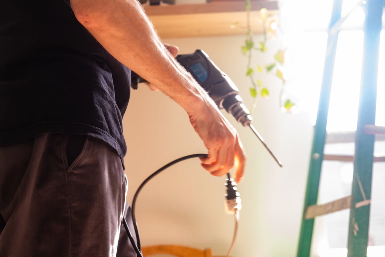 A person holding a power drill and plugging it in, with sunlight streaming through a window and a green ladder in the background.