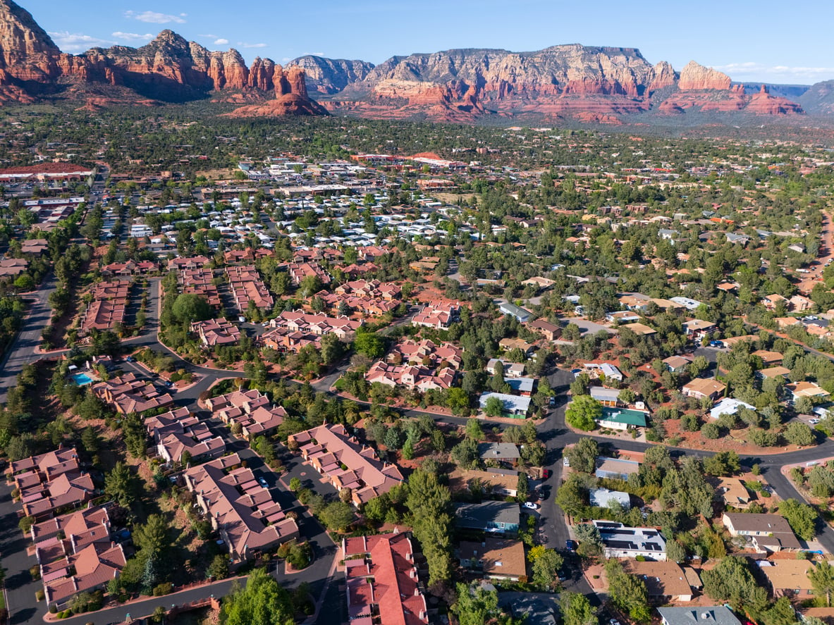 Aerial view of a suburban neighborhood with red-roofed houses, surrounded by green trees and set against red rock mountains under a blue sky—a picturesque scene that feels like one of the region’s hidden vacation spots.