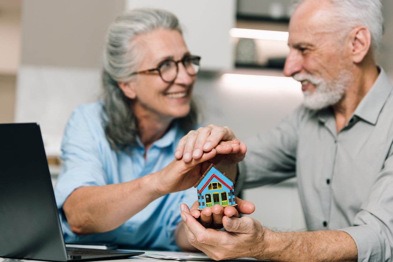 Smiling older couple sitting at a table, holding a small model house together, with a laptop in front of them, suggesting planning or discussing real estate or home ownership.