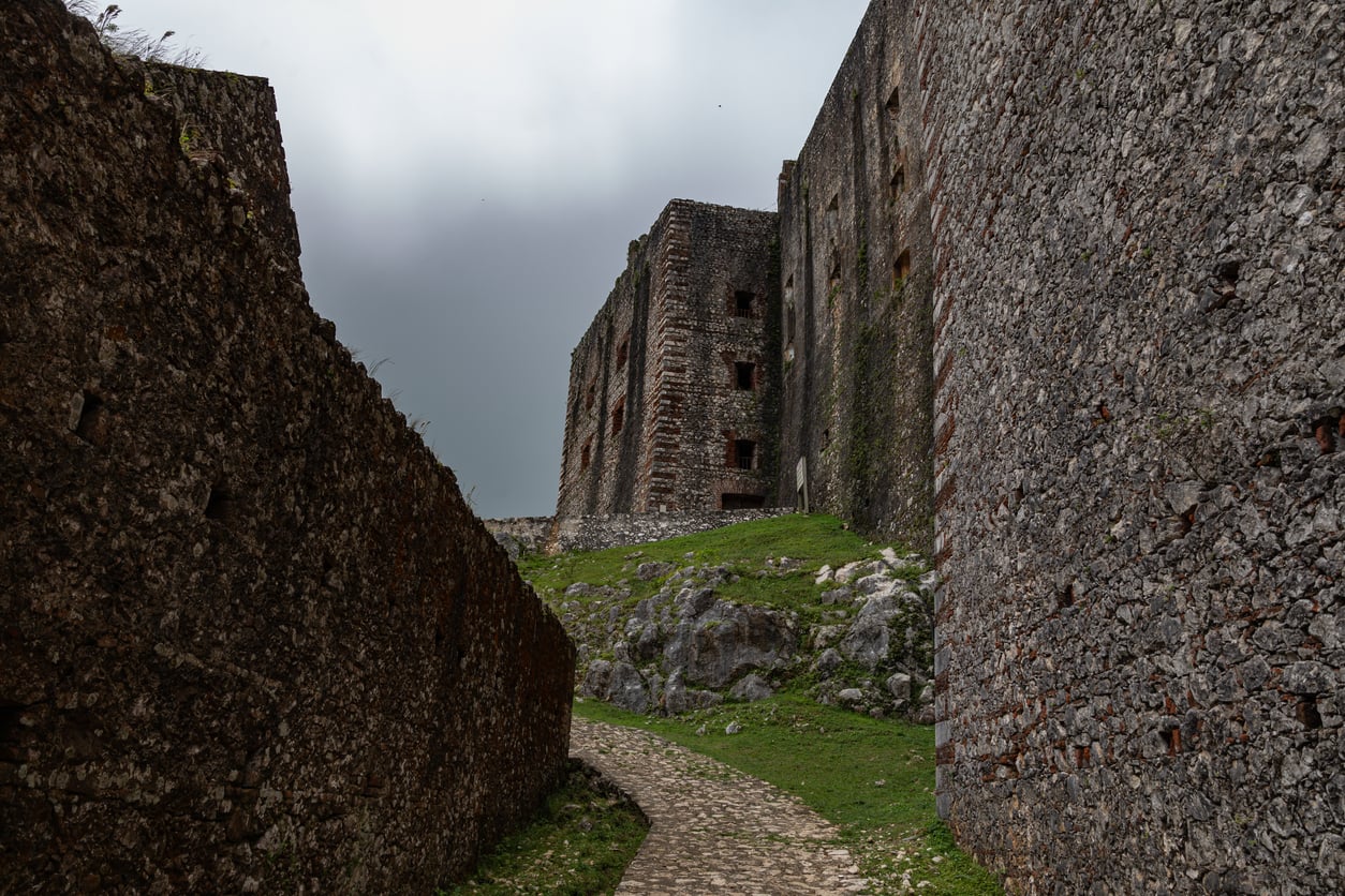 A narrow stone pathway leads between tall, weathered stone walls toward a large, historic fortress under a cloudy sky, with patches of green grass and rocks along the ground—a scene reminiscent of the secluded sites often mentioned among the Places Travelers Are Warning Against Going In 2026.