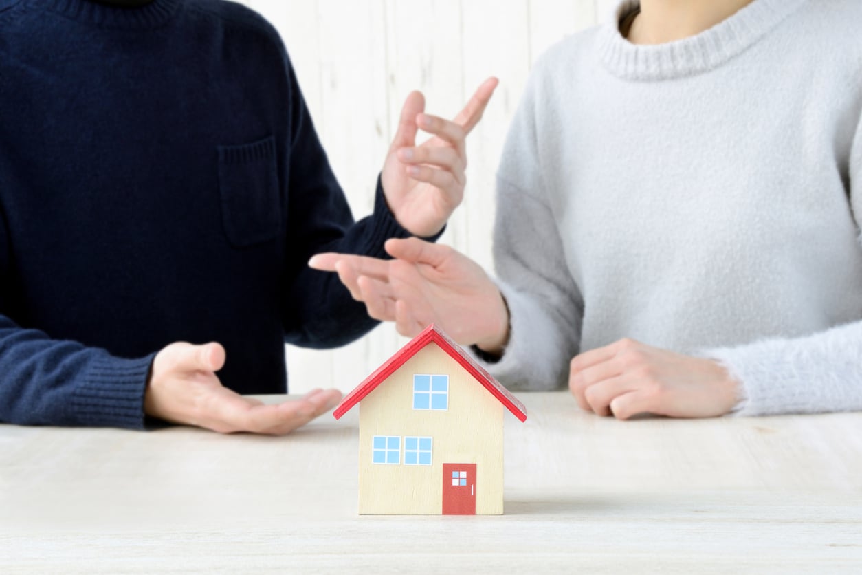 Two people gesture and appear to discuss something, with a small model house in the foreground on a table, suggesting a conversation about real estate or property.