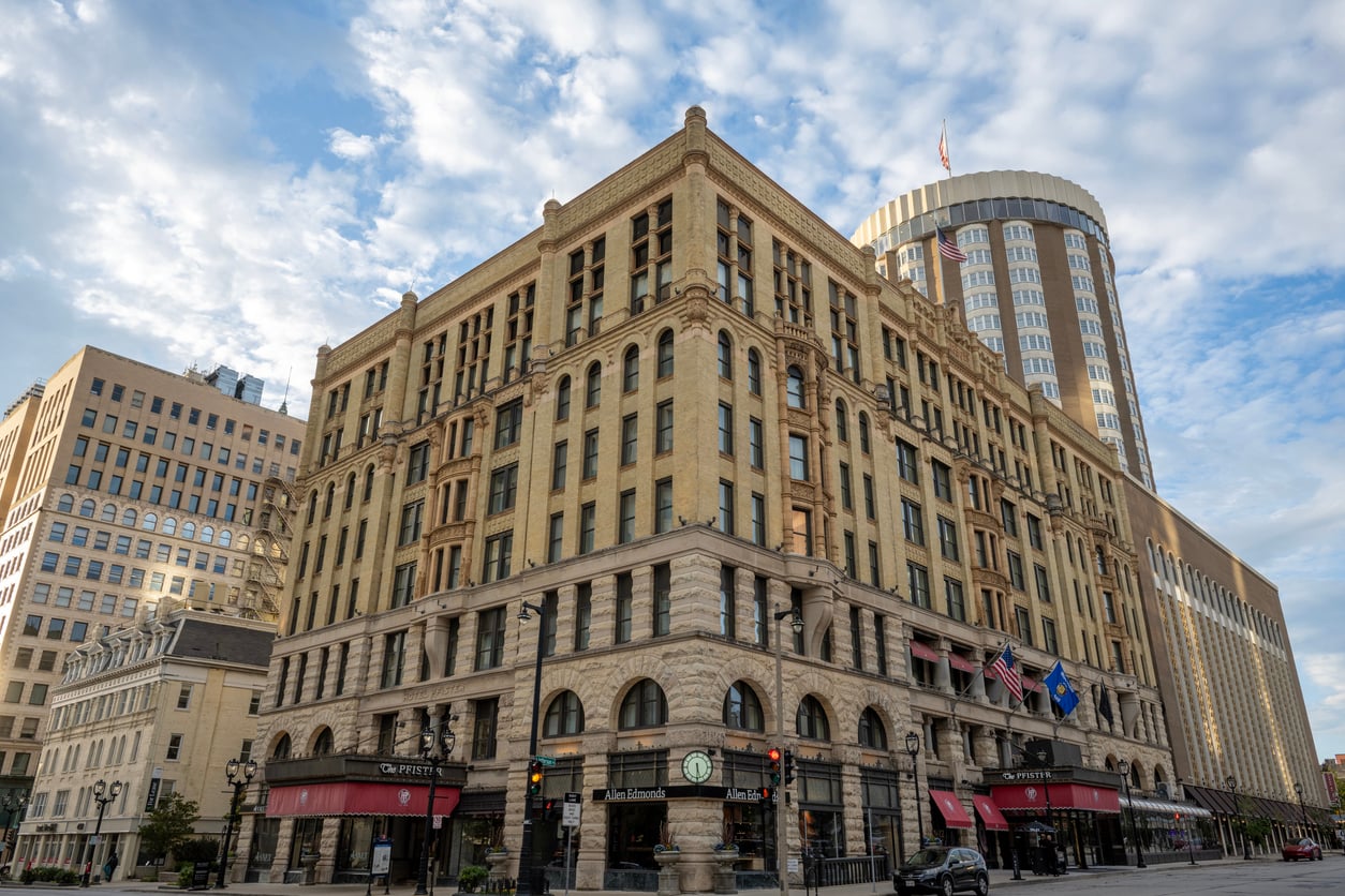 A large historic building, reminiscent of many renowned Historic Hotels, features a tan stone exterior, arched windows, and flags near the entrance. Behind it rises a tall round modern building. The sky is mostly blue with scattered clouds.