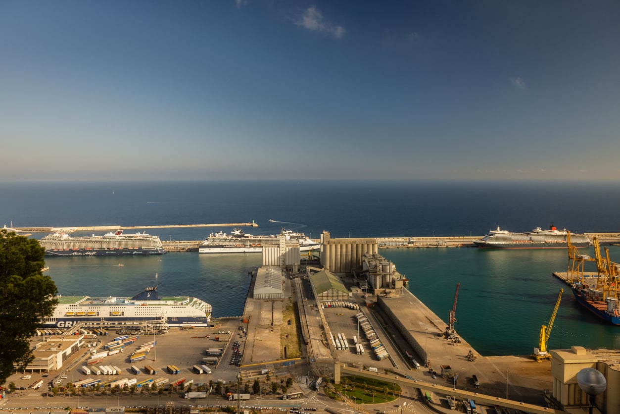 Aerial view of a large port with cruise ships docked, warehouses, cranes, and trucks. The sea is calm and blue, stretching to the horizon under a clear sky—a striking scene that contrasts sharply with recent discussions about Places Travelers Are Warning Against Going In 2026.