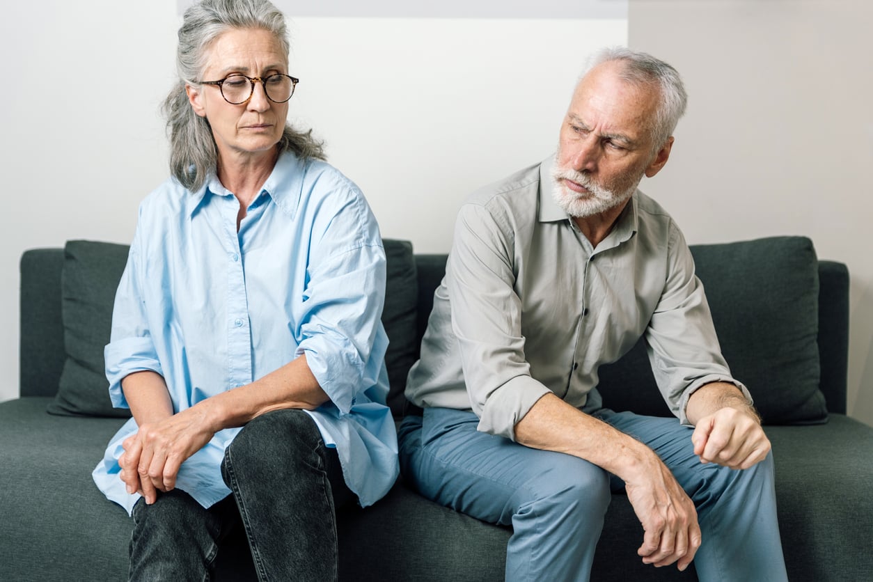 An older woman and man sit on a couch facing away from each other, both looking upset and avoiding eye contact. The room is neutral and their body language suggests tension or disagreement, perhaps highlighting the importance of communication and retirement tips for navigating this stage of life together.