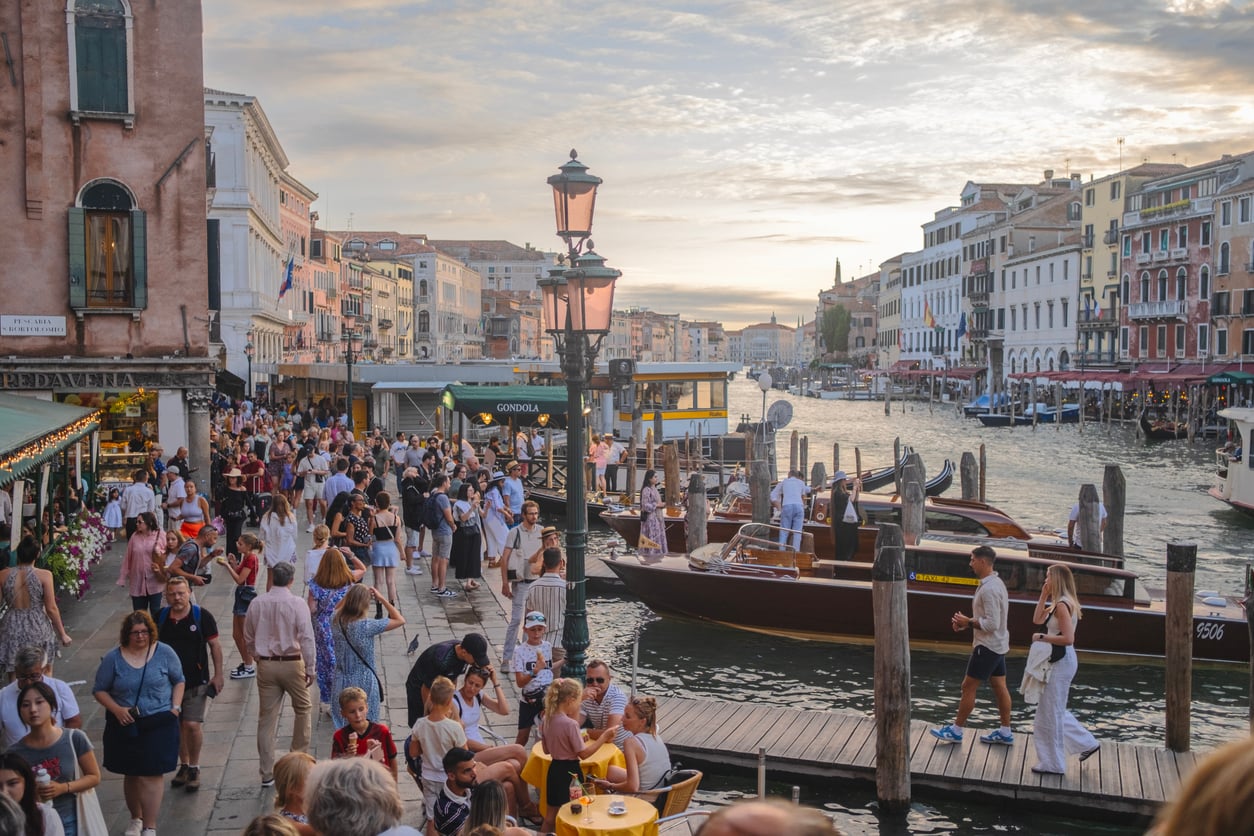 A crowded Venice canal scene at sunset with people walking, sitting, and boarding boats. Historic buildings line the water, and gondolas and water taxis are docked along the busy waterfront.