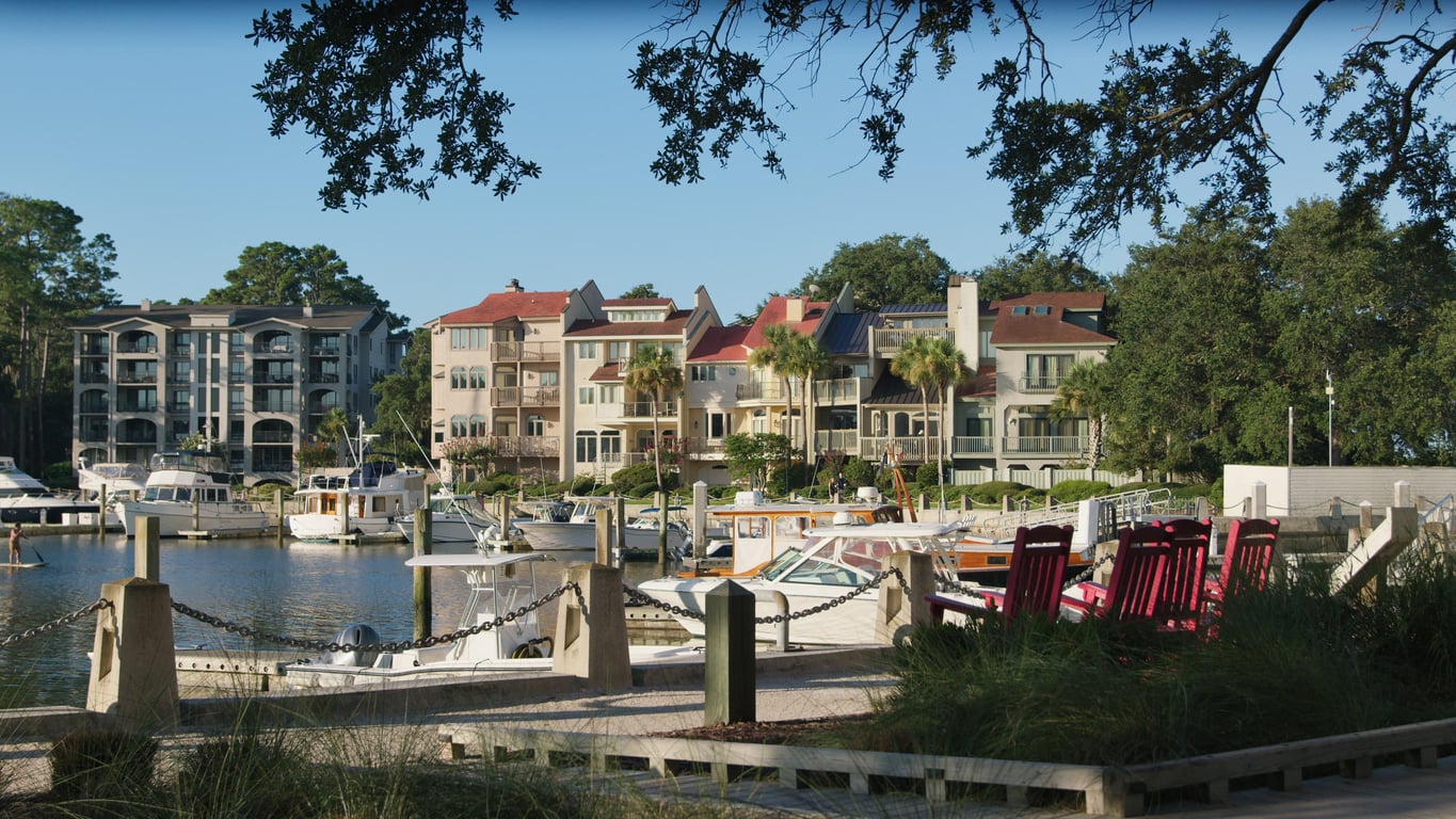A marina with docked boats, red Adirondack chairs in the foreground, and multi-story waterfront homes with balconies and palm trees in the background, framed by leafy tree branches—an inviting scene that captures the charm of top vacation spots.