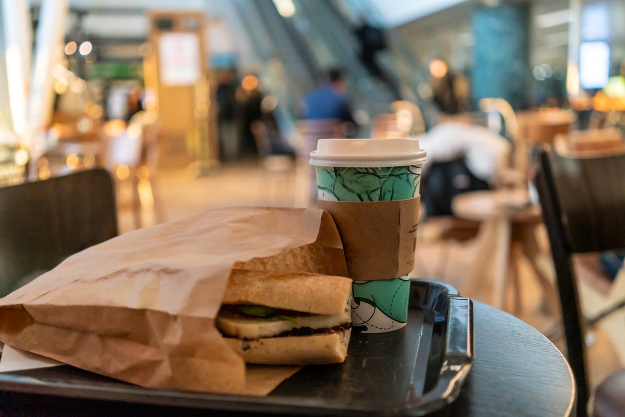 A sandwich partially wrapped in brown paper and a to-go coffee cup sit on a tray on a café table, with blurred people and tables in the background.